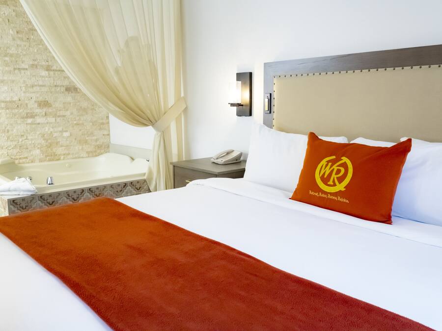 Een hotelkamer met een groot bed, een hoofdbord met een logo, een nachtkastje met een telefoon, een wandlamp en een bad.