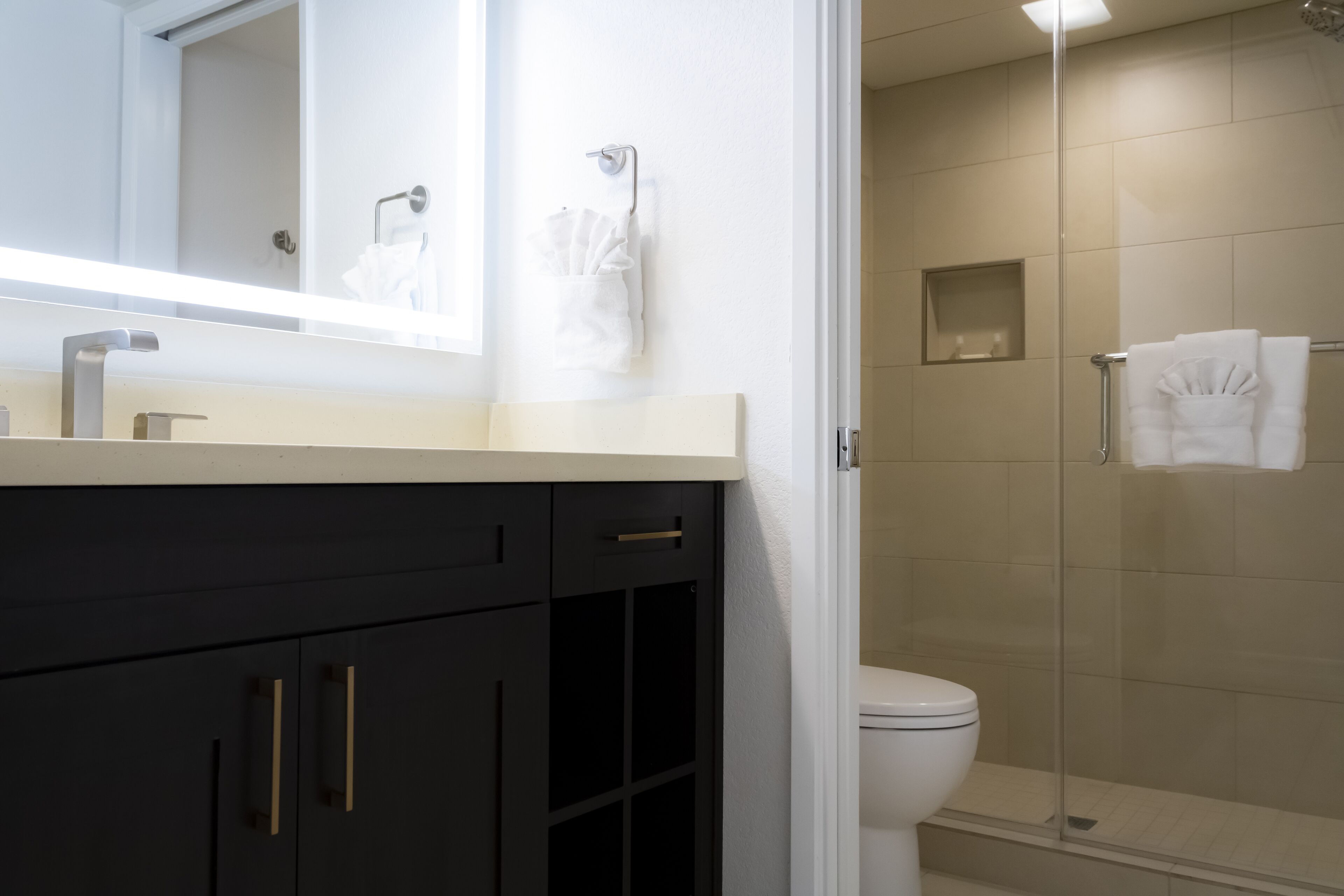 Un baño moderno con un mueble de tocador oscuro, un lavamanos blanco, una cabina de ducha con mampara de cristal y un inodoro blanco.