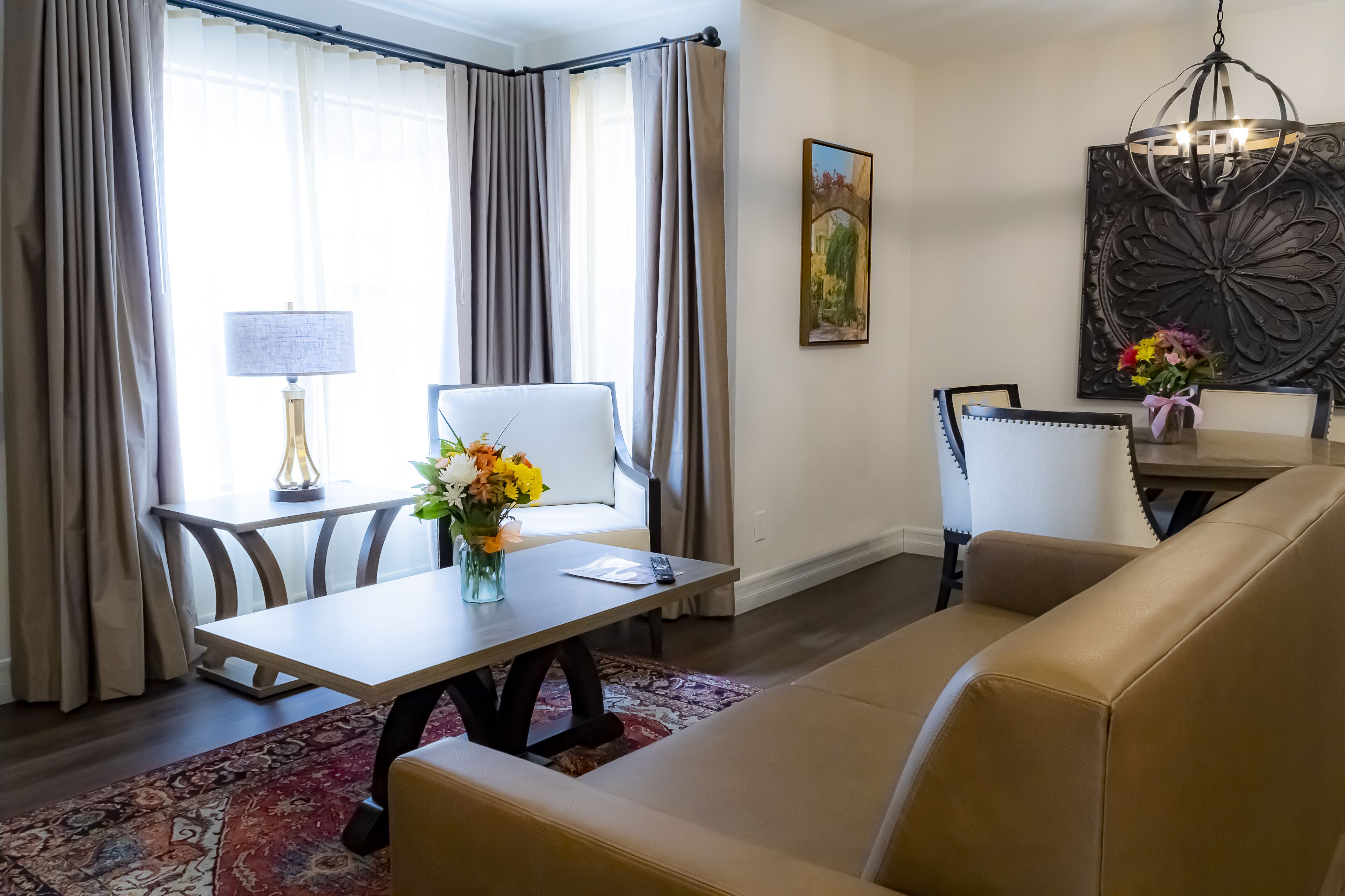 A modern living room with a beige sofa, a dining table with a vase of flowers, a chandelier, and a painting on the wall.
