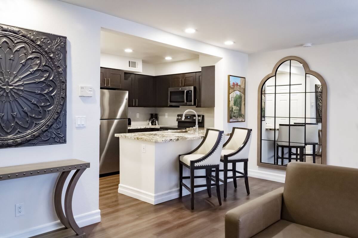 A modern kitchen with dark cabinetry, a stainless steel refrigerator, and a microwave. There is a small island with two chairs and a coffee maker.