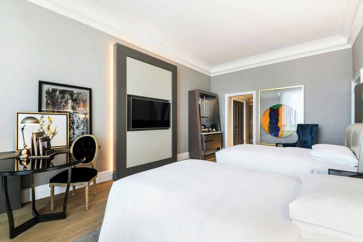 A hotel room with two beds, a desk with a chair, a TV, and artwork on the walls.