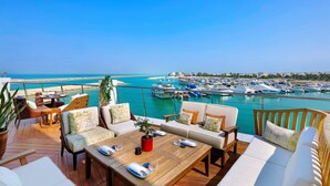 Una terraza con piso de madera, mobiliario exterior y vista a un puerto deportivo con barcos.