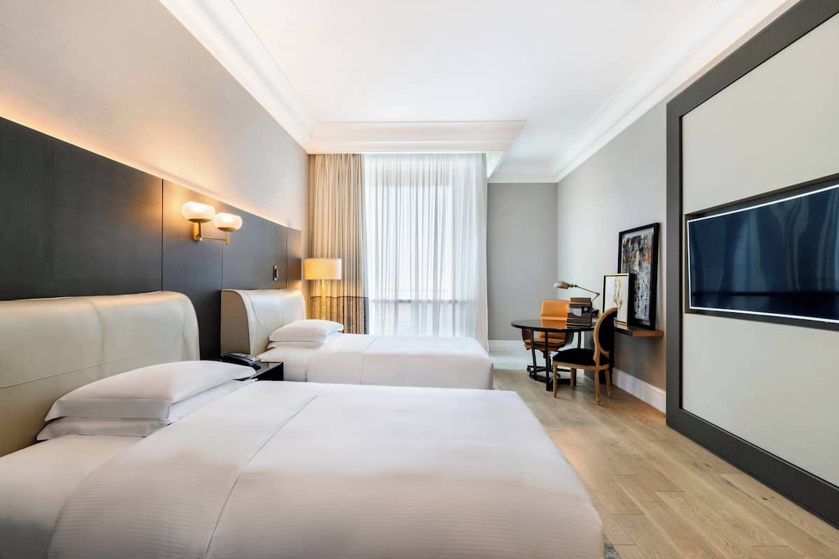 A hotel room with two beds, a desk with a chair, a television, and a large window with curtains.