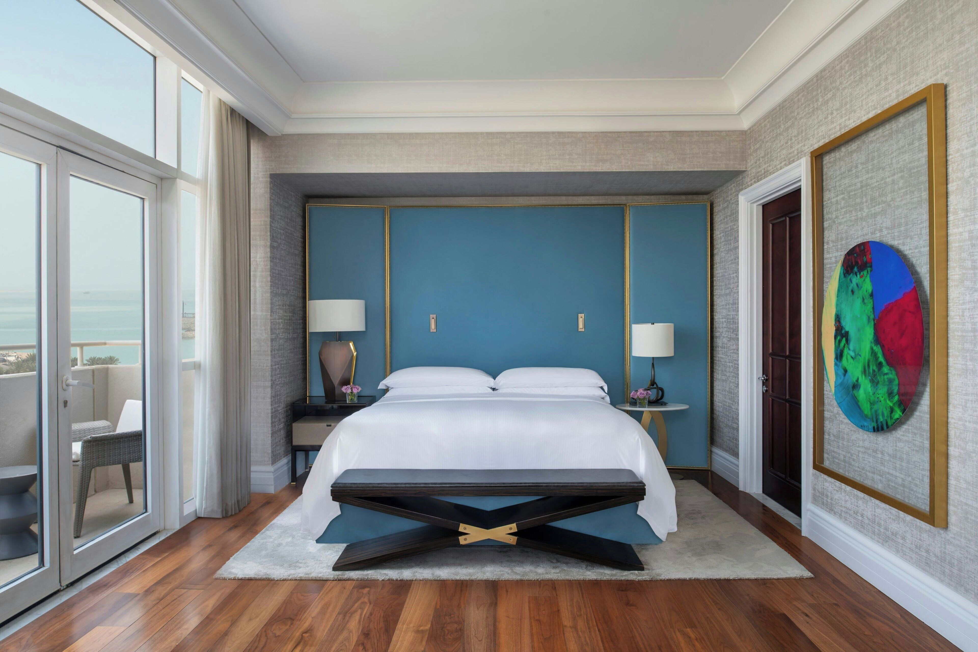 A spacious bedroom with a large bed, two bedside tables with lamps, a view of the outdoors through a sliding door, and a colorful abstract painting on the wall.