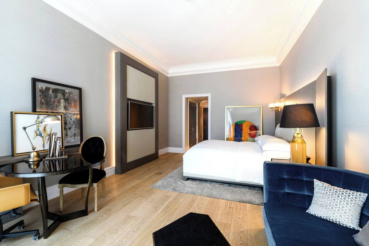 A modern hotel room with a bed, a desk with a chair, a sofa, and a TV mounted on the wall.