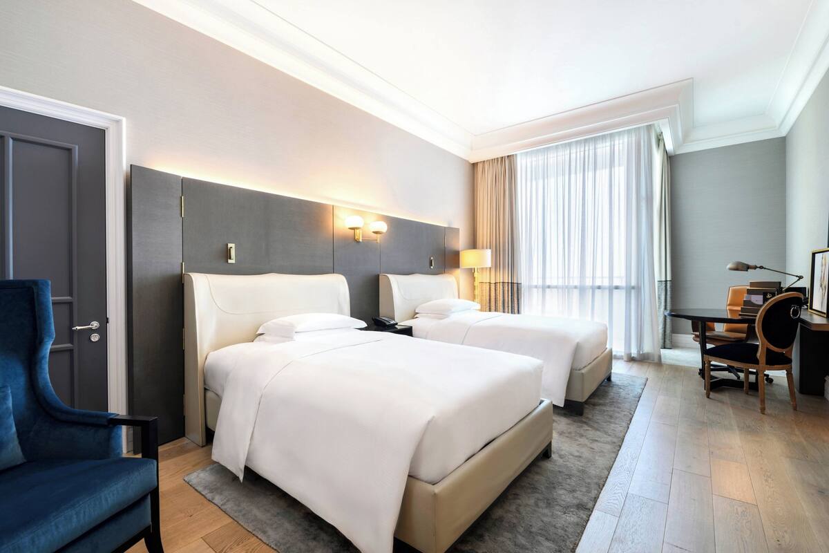 A hotel room with two beds, a desk, a chair, and a large window with curtains.