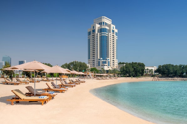A beach with sun loungers and umbrellas, a tall building in the background, and clear blue water.