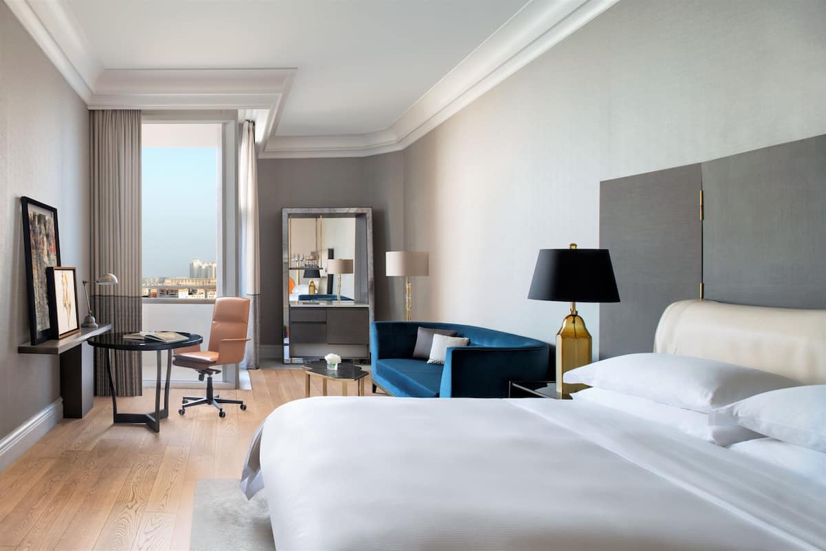 A hotel room with a large bed, a blue sofa, a desk with a chair, a lamp, and a view of the city.