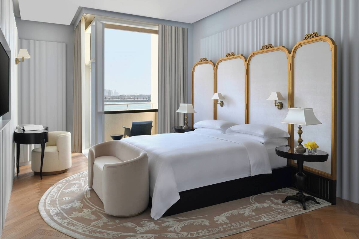 A hotel room with a large bed, two bedside tables with lamps, a chair, a small table, and a view of the city.
