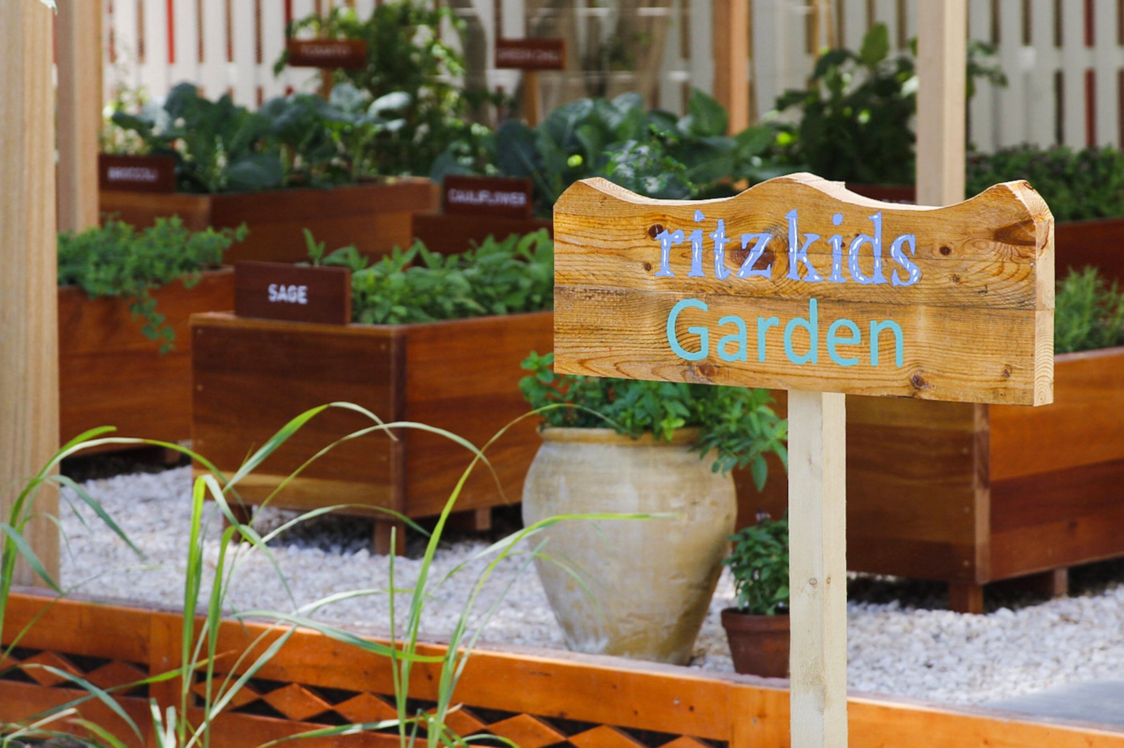 Un letrero de madera con el texto 'Ritzkids Garden' en un jardín con diversas plantas y macetas de madera.