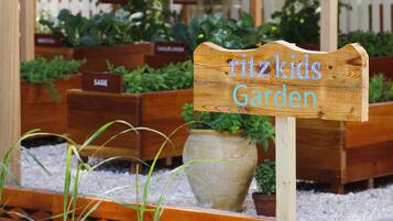 Un letrero de madera con el texto 'Ritzkids Garden' en un jardín con diversas plantas y macetas de madera.