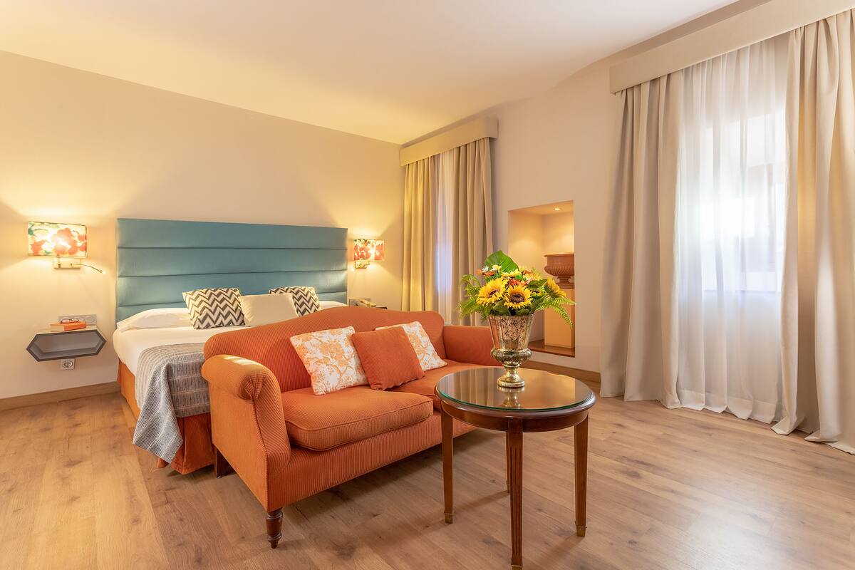 A hotel room with a bed, a sofa, a small table with a vase of flowers, and a window with curtains.