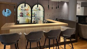 A modern bar area with a wooden counter, three bar stools, and a glass display of bottles. - Hotel Langwieder See (Munich)