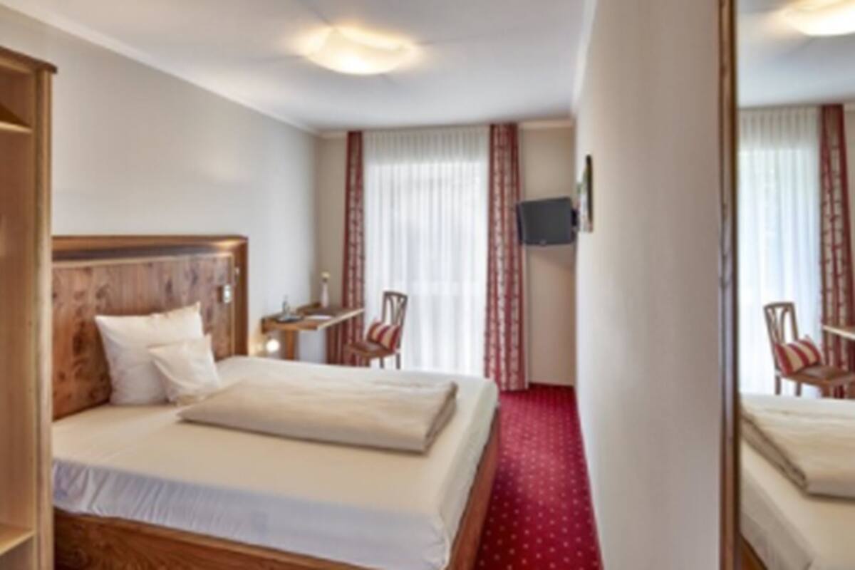 Habitación de hotel con cama de cabecera de madera, televisor de pantalla plana montado en la pared, un pequeño escritorio con silla y suelo cubierto de alfombra roja.