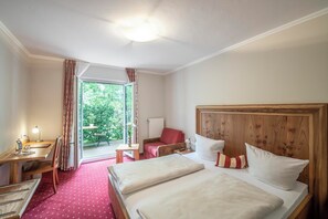 A hotel room with a wooden headboard, two beds, a desk with a lamp, a chair, and a view of greenery through the window. - Hotel Langwieder See (Munich)