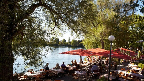 An outdoor dining area with tables and chairs, overlooking a body of water, surrounded by trees. - Hotel Langwieder See (Munich)