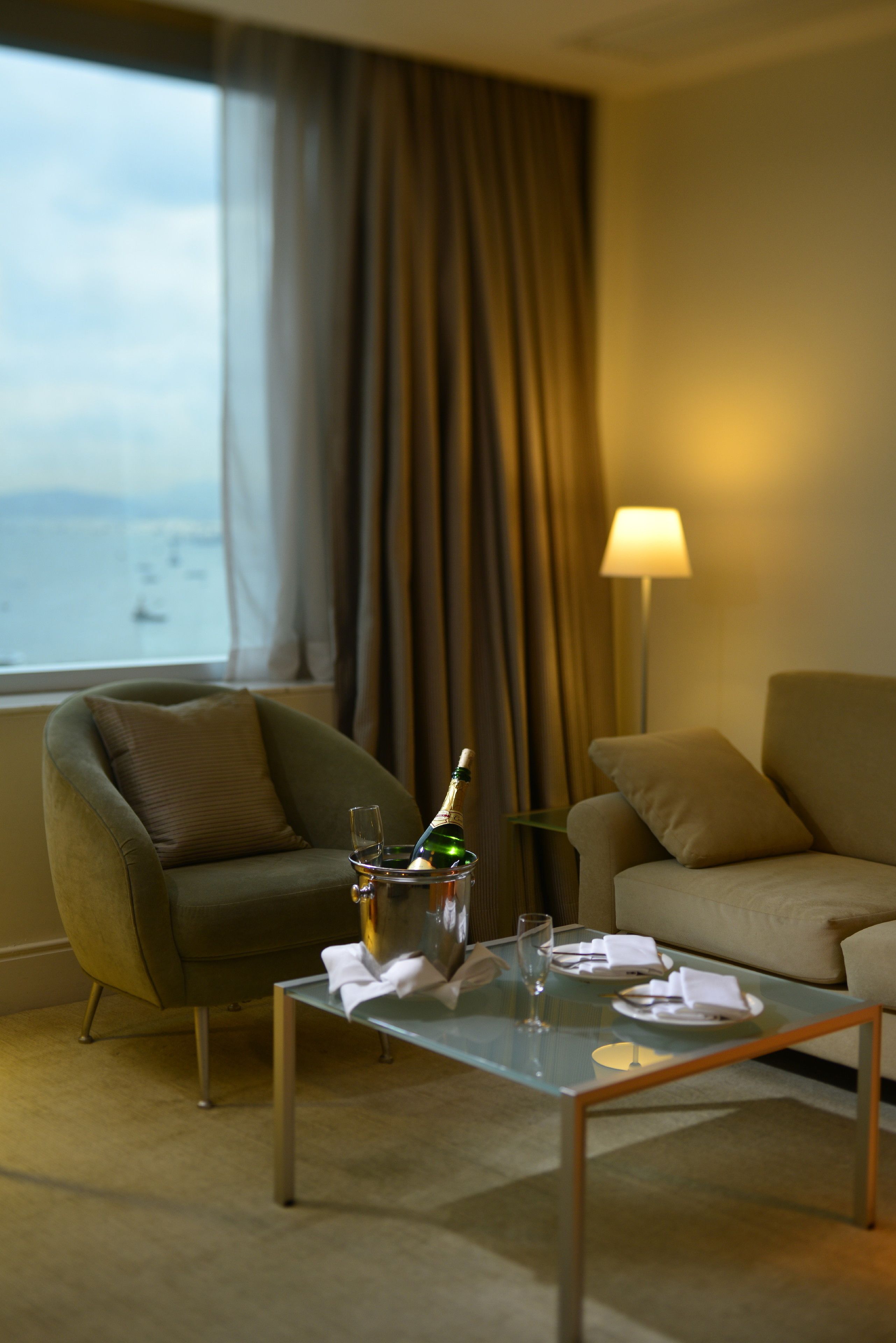 A hotel room with a view of the sea, a green armchair, a beige sofa, a glass coffee table with a bottle and glasses, and a lamp.