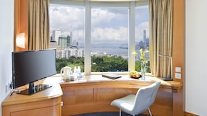 Hotelkamer met een groot raam met uitzicht op de stad, een bureau, een flatscreen-tv, een stoel en een vaas met bloemen.