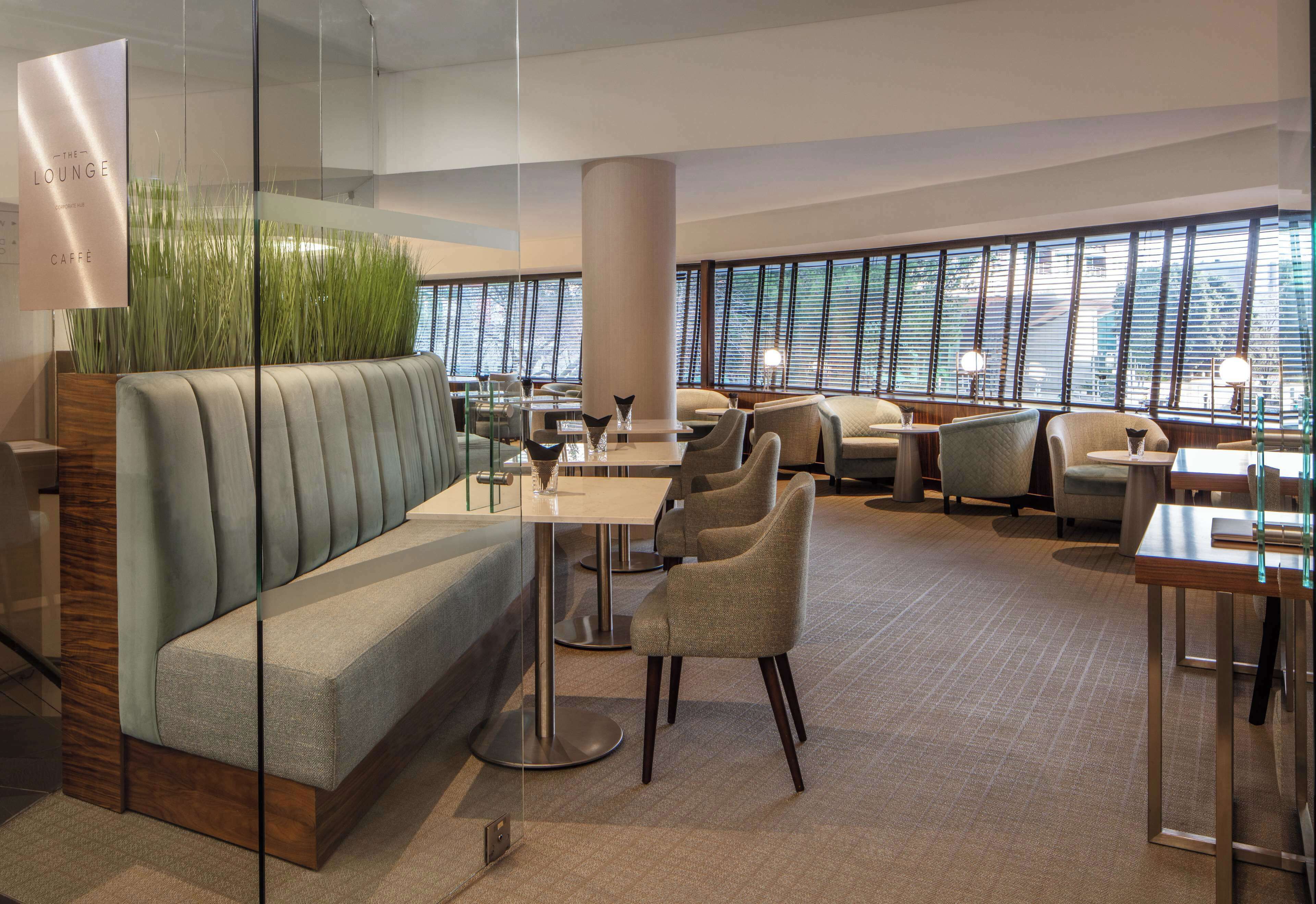 A modern lounge with a sofa, chairs, tables, and a large window with blinds.