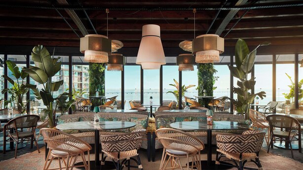 Un restaurante con mesas redondas y sillas de mimbre, grandes lámparas colgantes y amplias ventanas con vistas al mar.