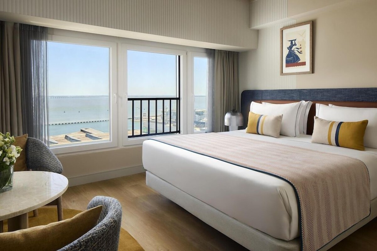 Habitación de hotel con una cama grande, una mesa redonda y vistas al mar a través de amplias ventanas.