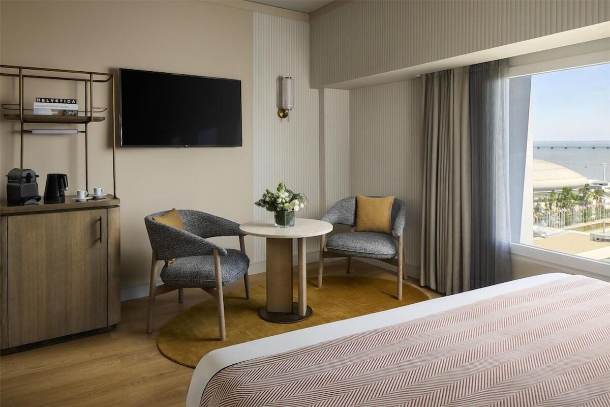 A hotel room with a bed, a desk with a coffee maker, two chairs, a TV, and a view of the city.