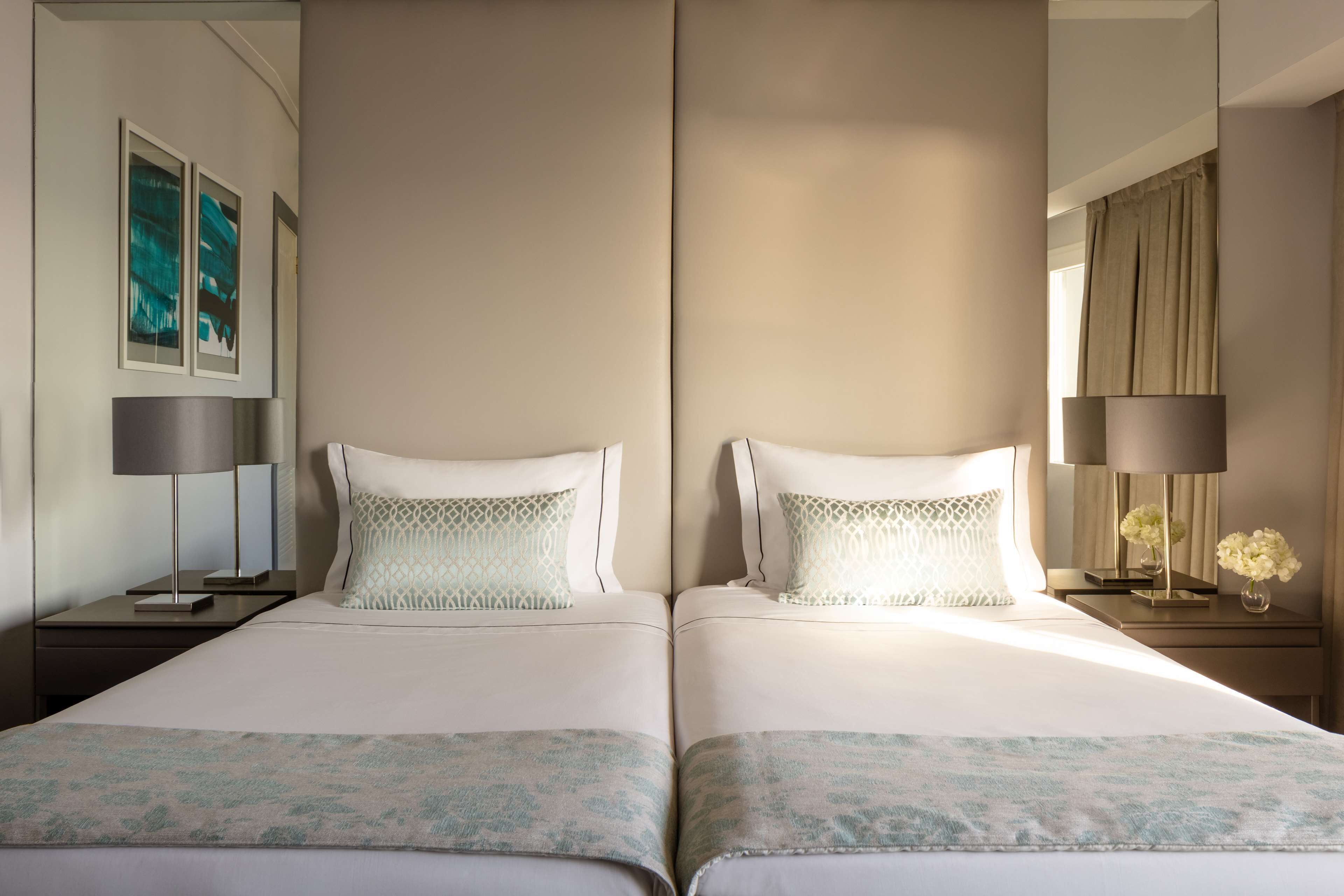 A hotel room with two beds, a nightstand with a lamp, a mirror, and artwork on the wall.