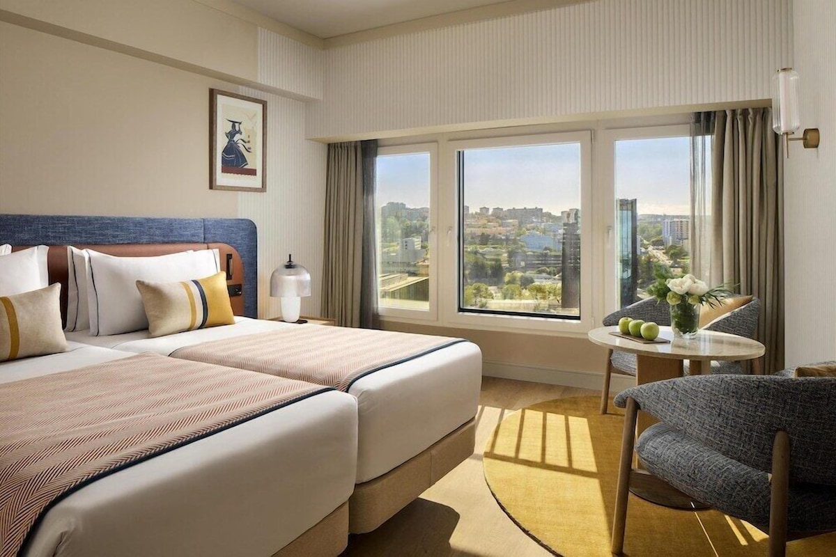 A hotel room with two beds, a small table with a vase, a chair, and a large window with a city view.
