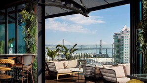 Un espacio exterior moderno con mobiliario de mimbre, suelo de madera y vistas a un puente y al paisaje urbano.