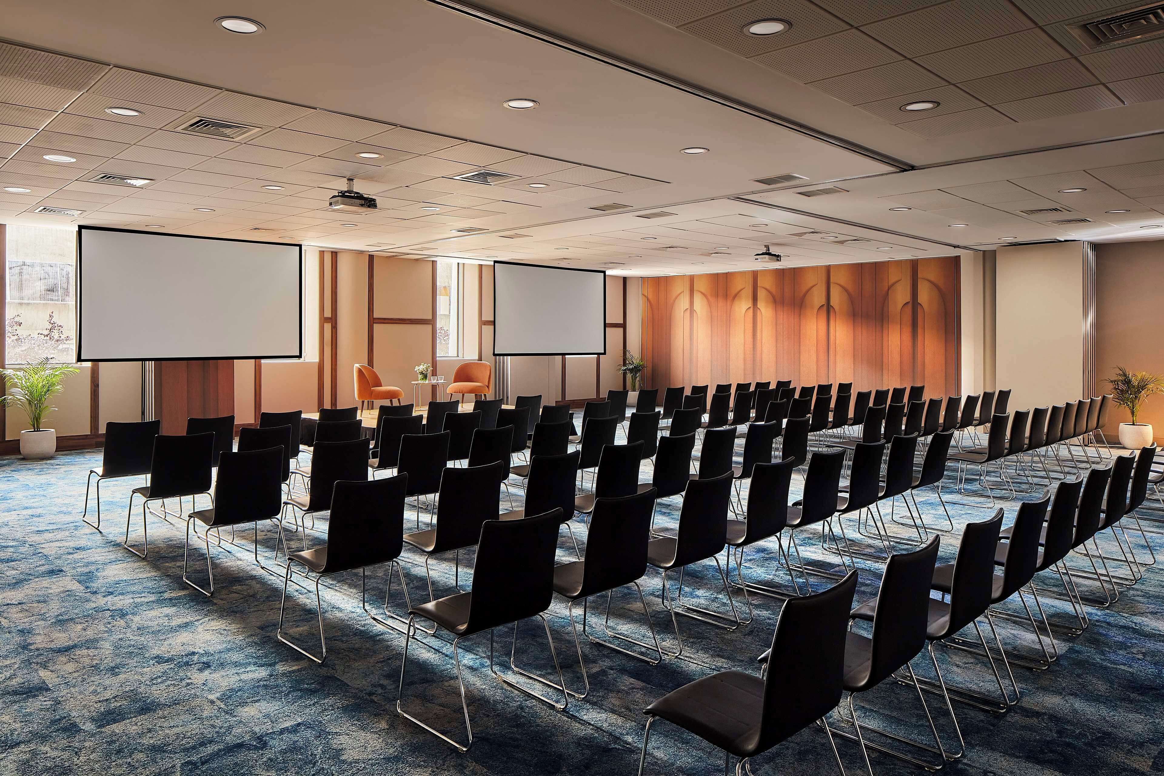 Un salón de conferencias con filas de sillas negras, una pantalla grande y piso alfombrado.