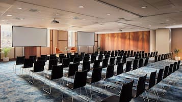Un salón de conferencias con filas de sillas negras, una pantalla grande y piso alfombrado.