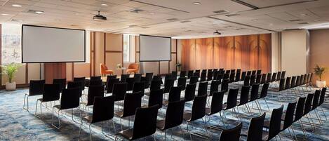 Un salón de conferencias con filas de sillas negras, una pantalla grande y piso alfombrado.