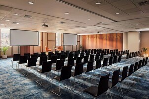 Konferenssihuone, jossa on riveissä mustia tuoleja, suuri näyttö ja matolla peitetty lattia.