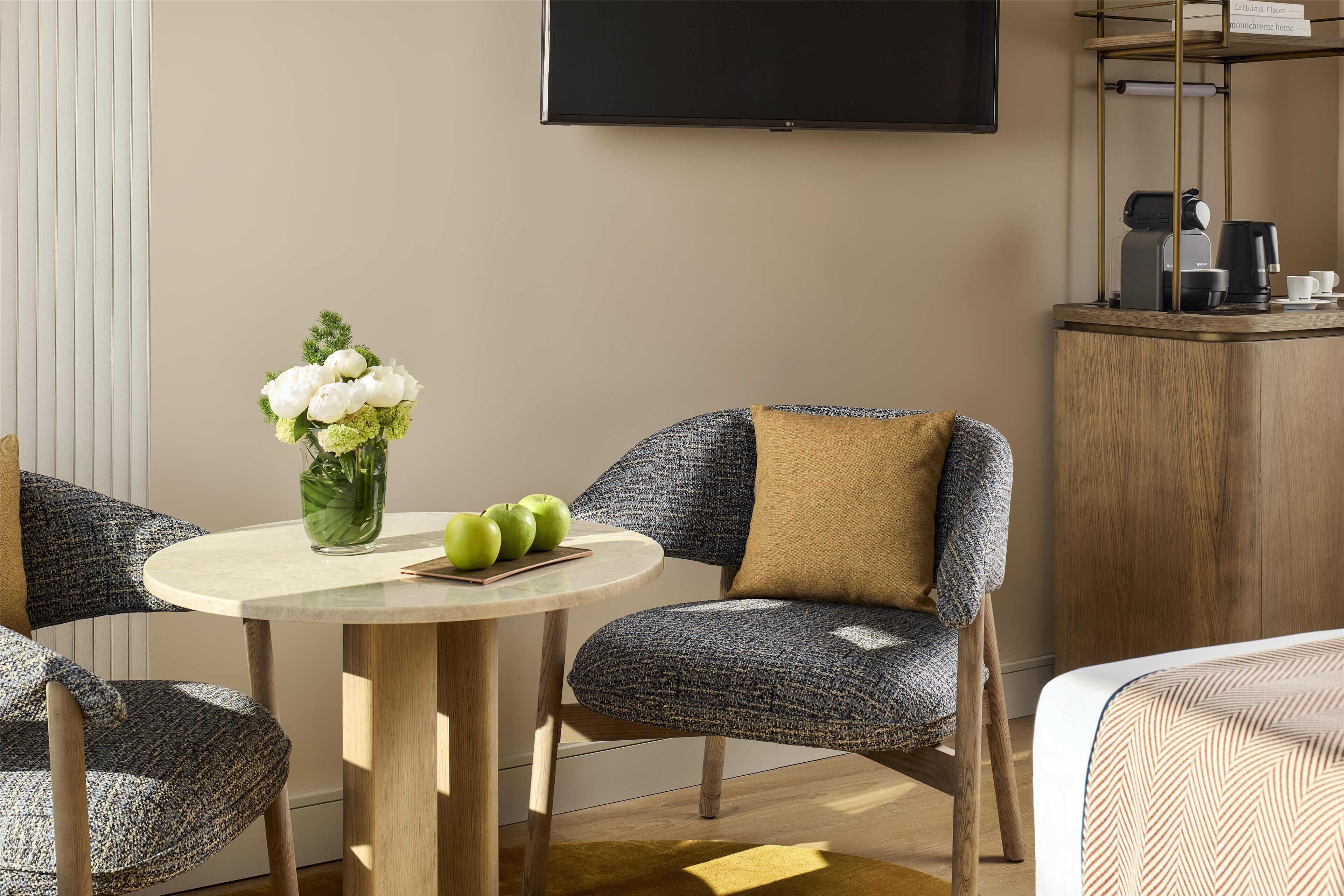 A hotel room with a round table, two chairs, a vase with flowers, and a coffee station.