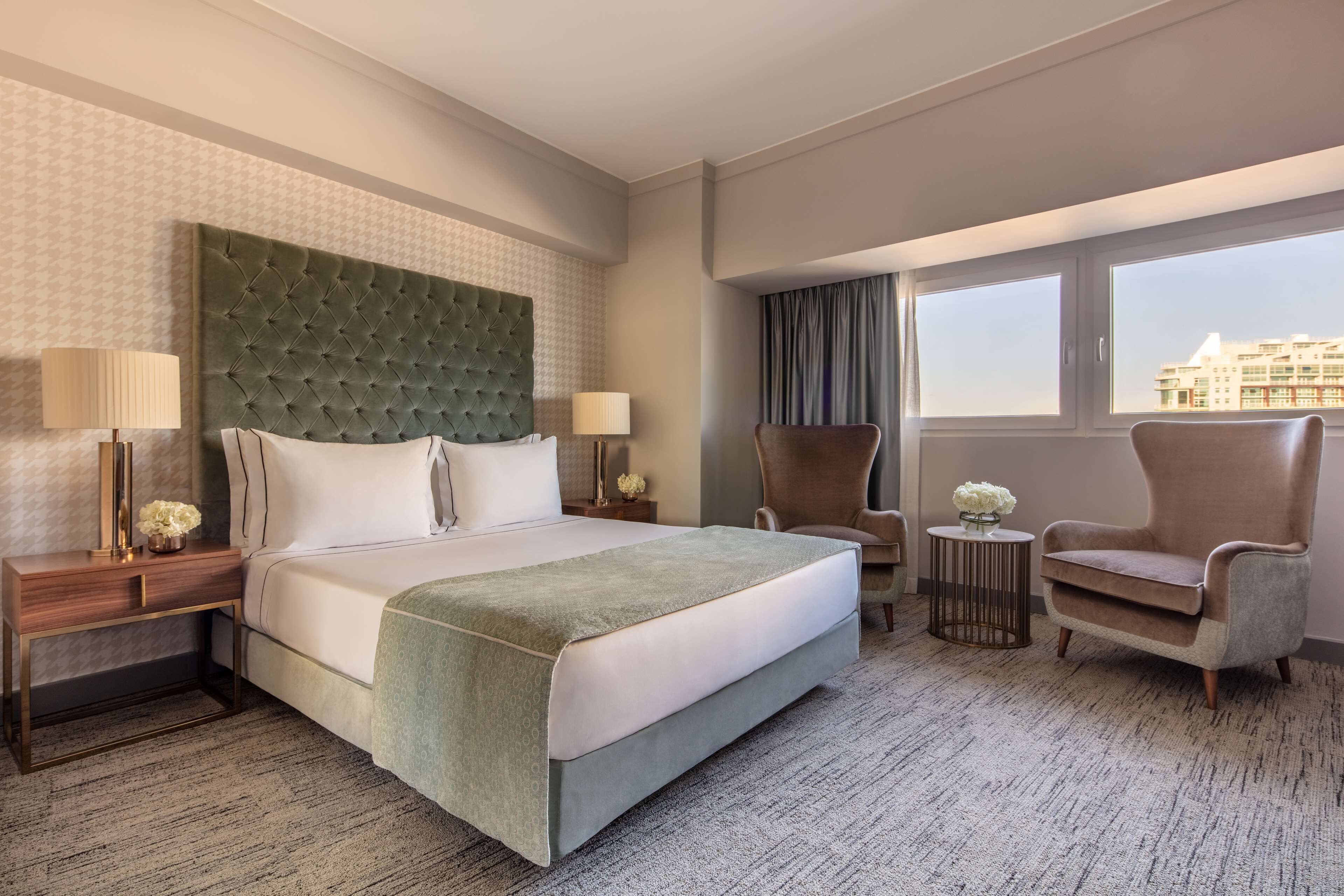 A hotel room with a large bed, two side tables, a chair, and a view of buildings through the window.