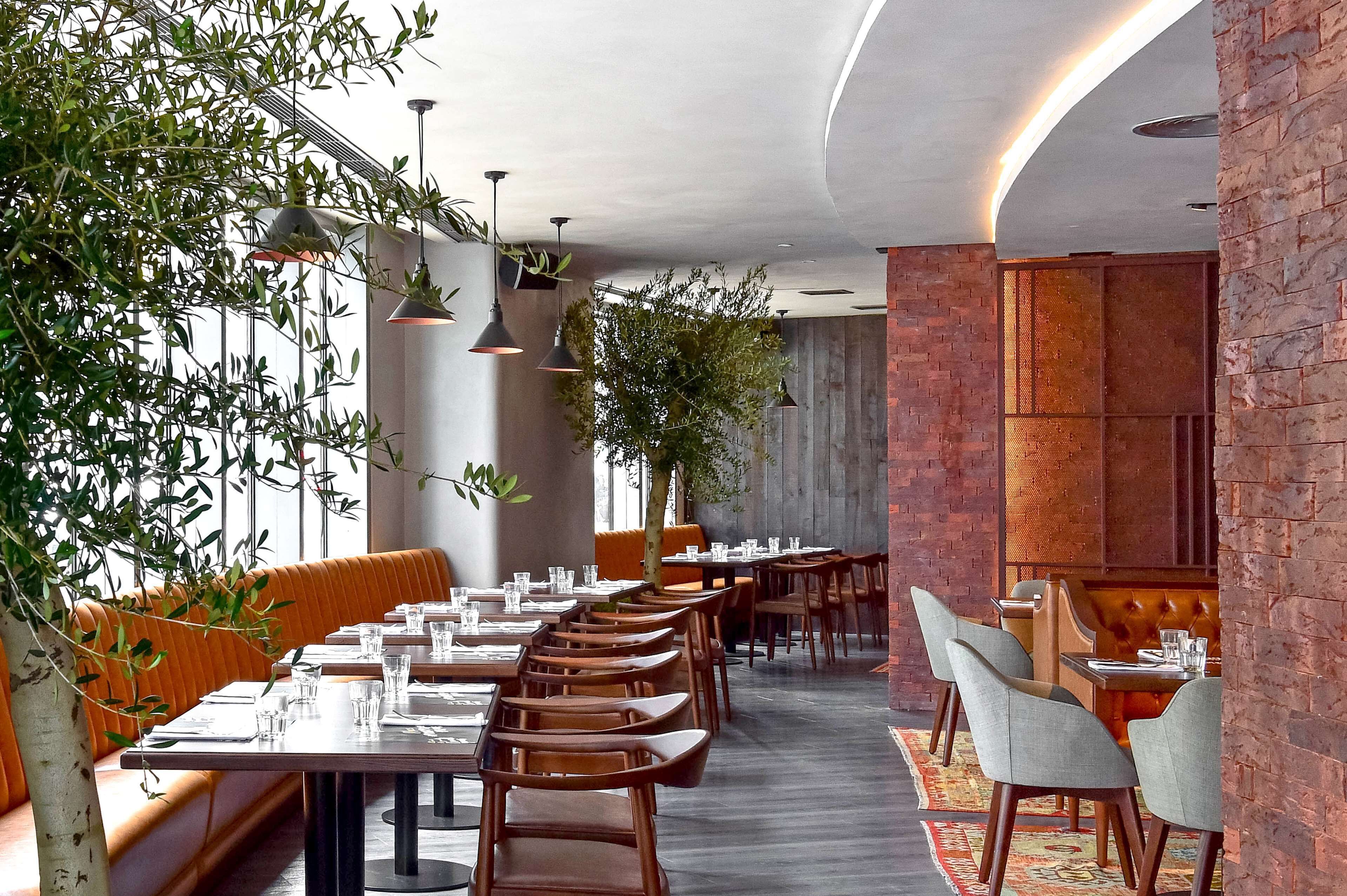 Un restaurante con largas mesas de madera, bancos con cojines naranja y lámparas colgantes.