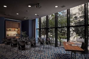 A conference room with a large table and chairs, a wooden cabinet with plants, and large windows offering a view of trees and buildings.
