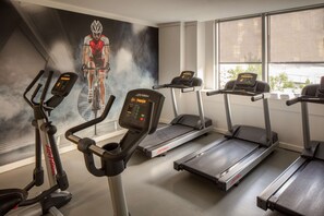 Une salle de sport équipée de tapis de course et de vélos d’appartement, ornée d’une fresque représentant un cycliste, et dotée de grandes fenêtres.