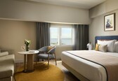 A hotel room with a bed, a chair, a small table with a vase, and a view of the sea through a window.