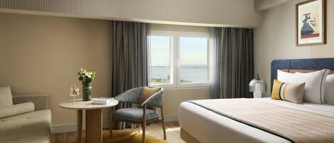 Habitación de hotel con cama, una silla, una mesita con un florero y vista al mar a través de una ventana.