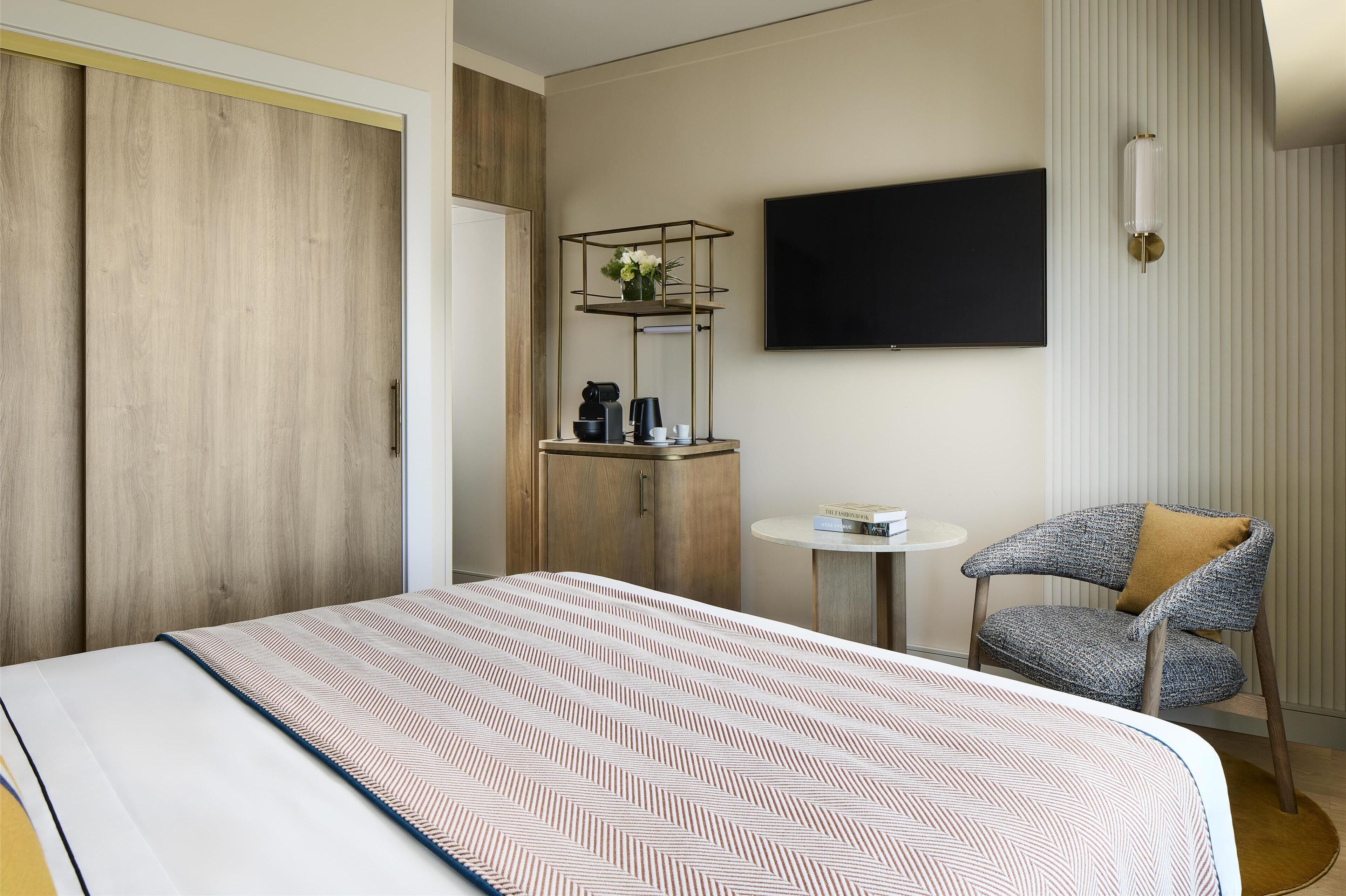 A hotel room with a bed, a chair, a small table, a TV, and a wardrobe.