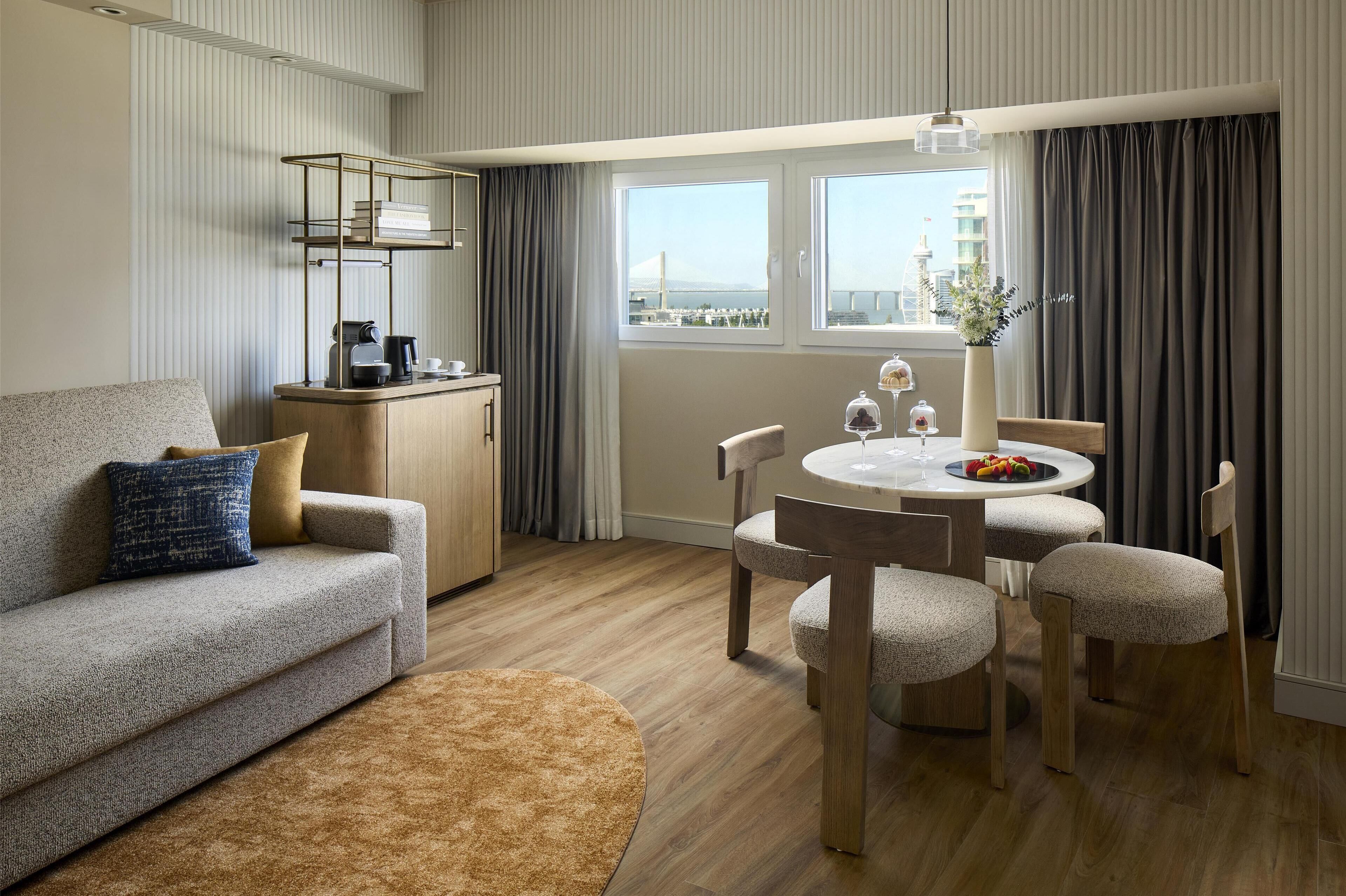 A modern hotel room with a sofa, a dining table with chairs, a small kitchen area, and a view of a bridge.