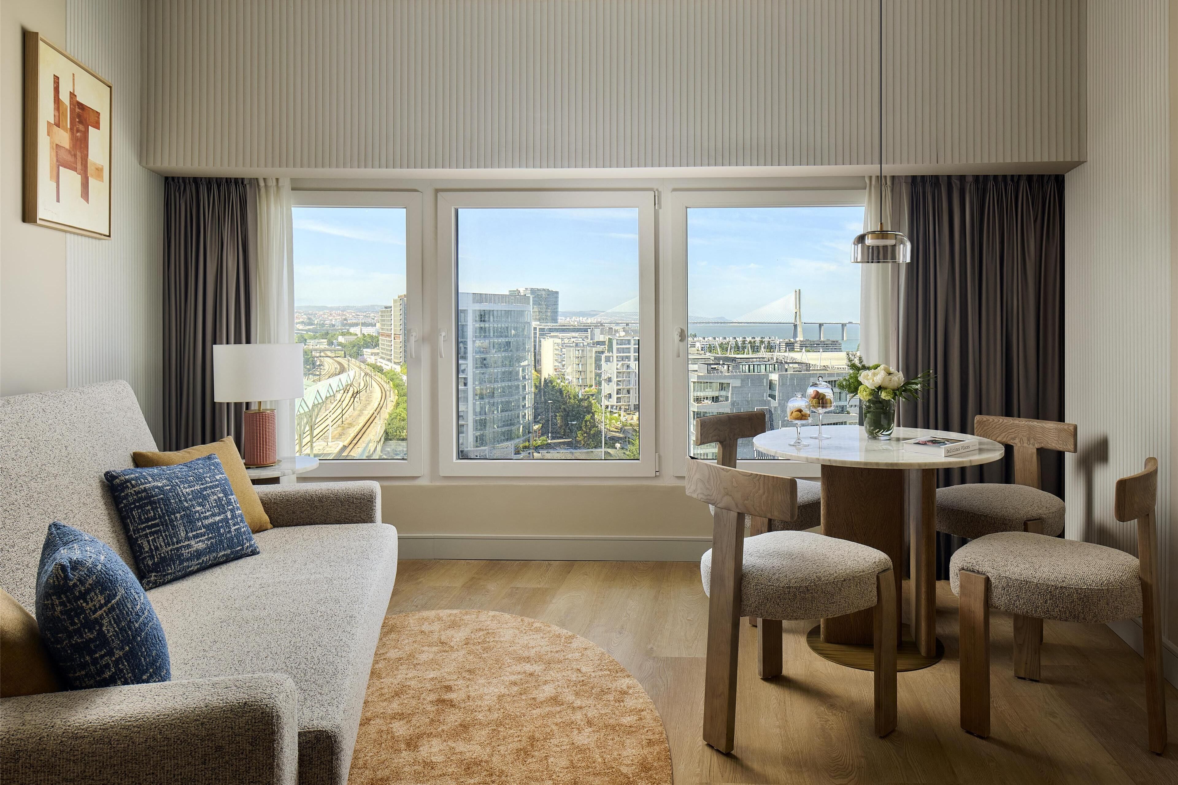 A modern living room with a sofa, a round table with chairs, and a large window offering a city view.
