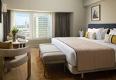 A hotel room with a large bed, a seating area with a chair and a small table, a view of the cityscape through the window, and a painting on the wall.
