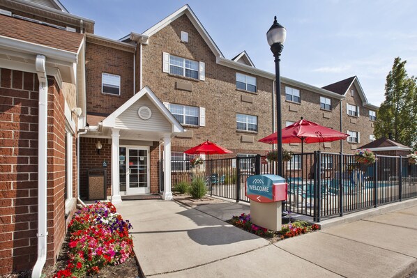 A brick building with a white entrance, red umbrellas, and a pool area. - TownePlace Suites Detroit Sterling Heights (Sterling Heights)