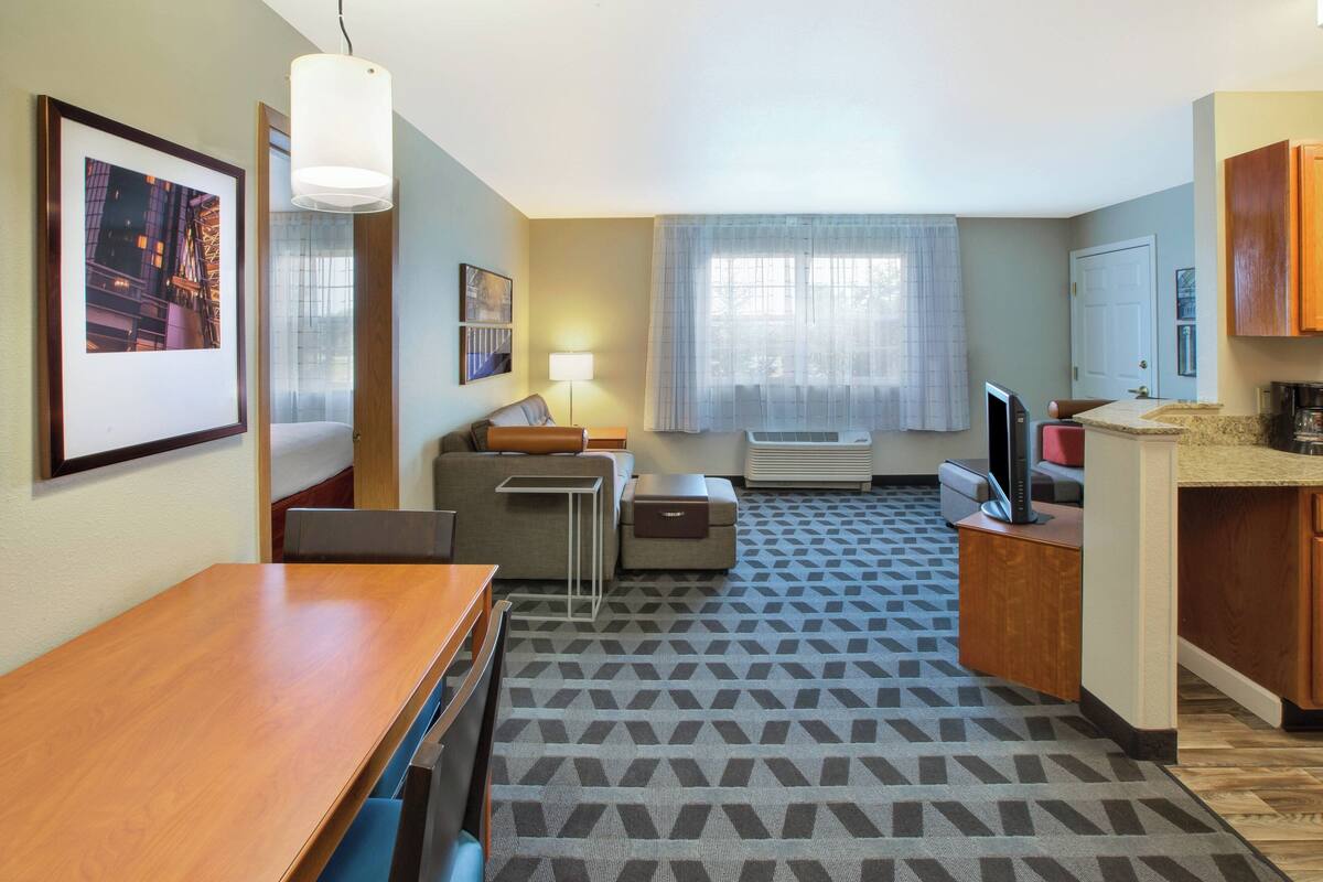 A hotel room with a wooden dining table, a sofa, a small side table, a TV, a mini-fridge, a microwave, and a window with sheer curtains.
