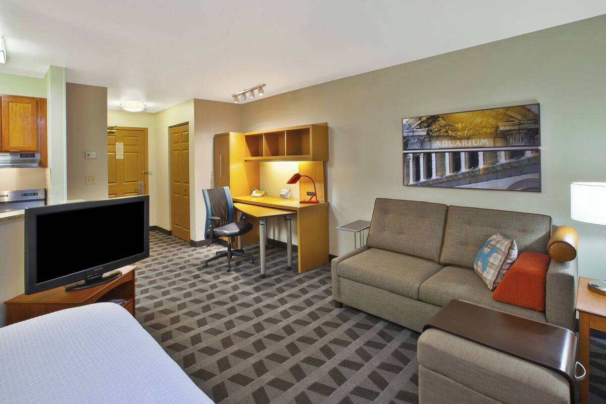 A hotel room with a bed, a TV, a desk with a chair, a sofa, and a painting of an aquarium.