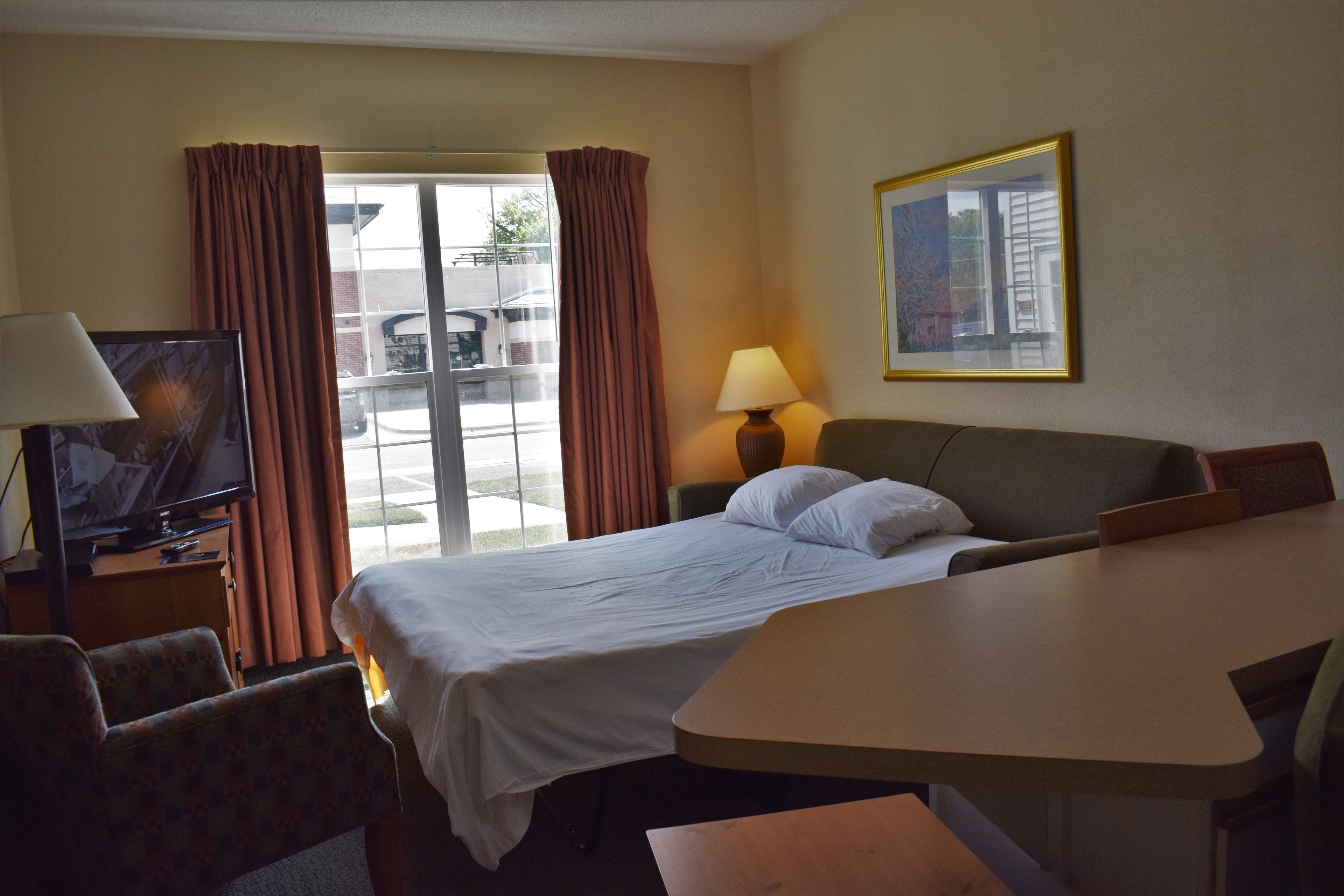Quarto de hotel com cama, escrivaninha, cadeira, televisão, abajur e janela com cortinas.
