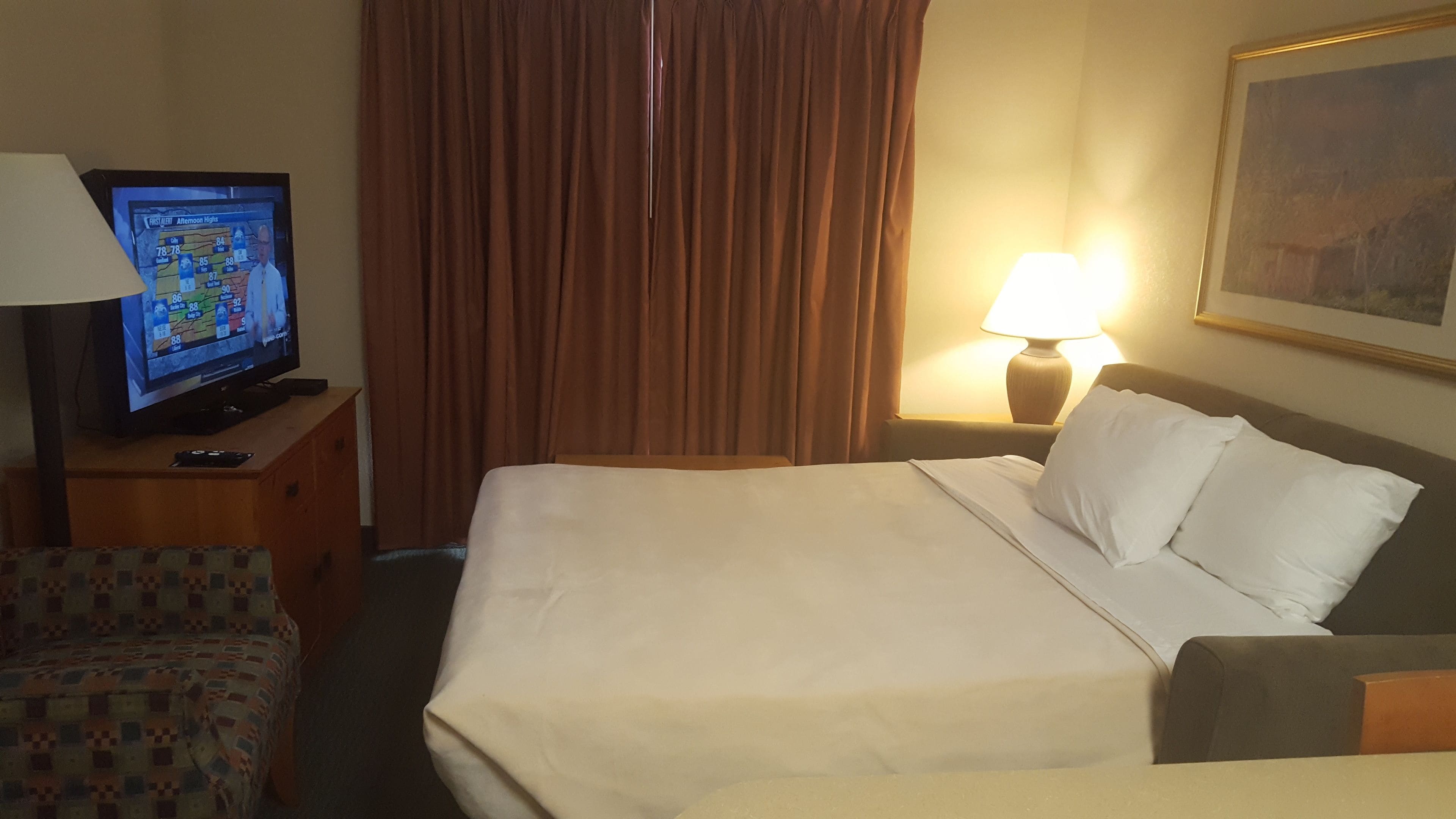 Ein Hotelzimmer mit einem Bett, einem Fernseher auf einem Nachttisch aus Holz, einer Lampe und einem Bild an der Wand.