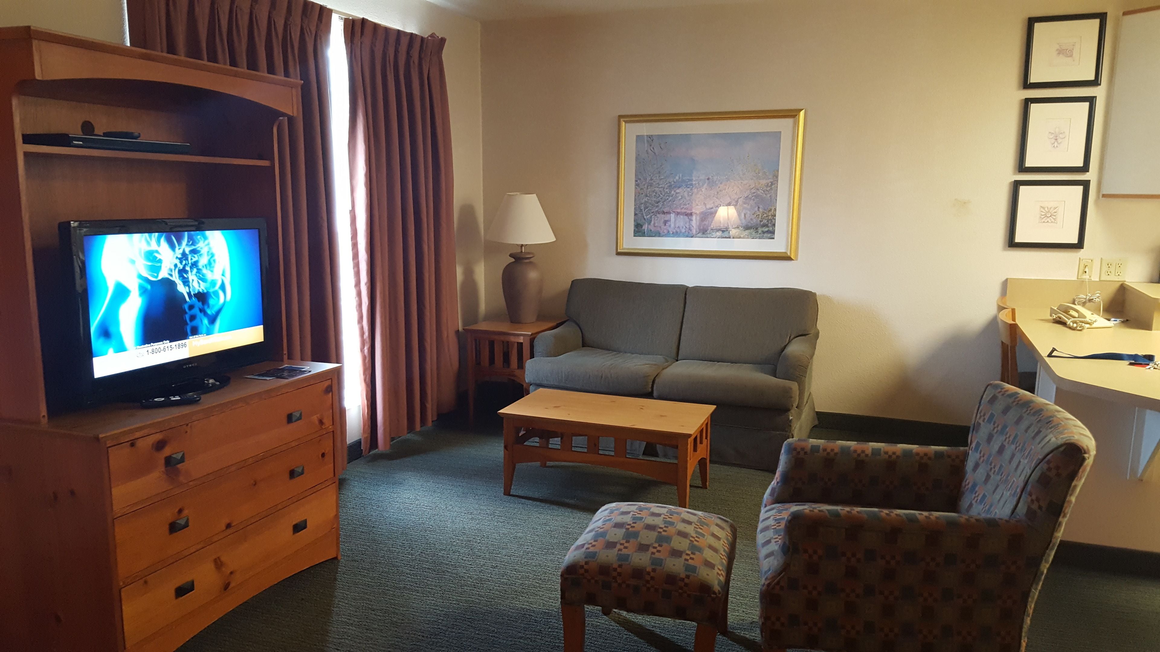 A hotel room with a television, sofa, armchair, wooden side table, and a desk with a lamp.